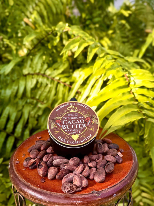 Theobroma Cacao Butter-Massage Candle placed on a wooden table with cacao beans and ferns in the background.