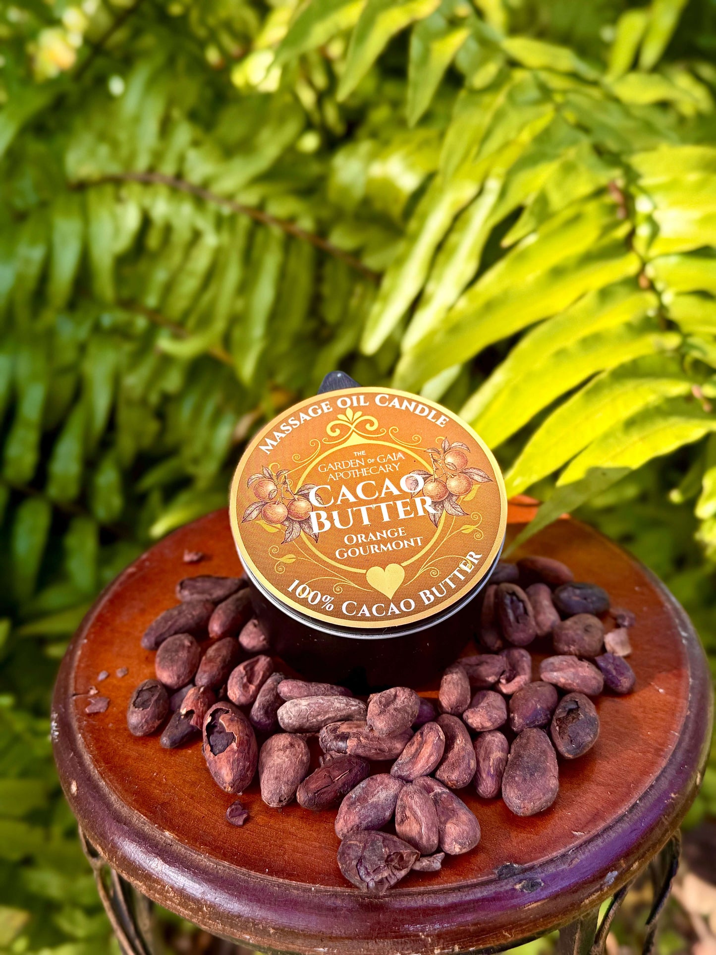 Theobroma Cacao Butter-Massage Candles-orange with cocoa beans on a wooden surface surrounded by green foliage.
