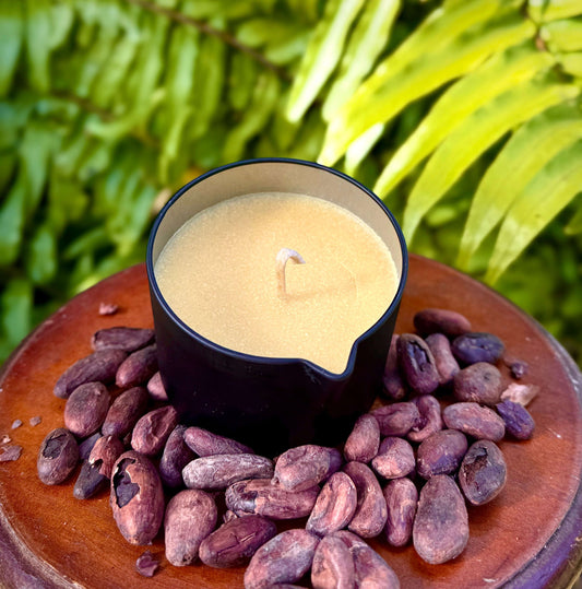 Theobroma Cacao Butter-Massage Candles-orange displayed on cocoa beans surrounded by lush greenery.