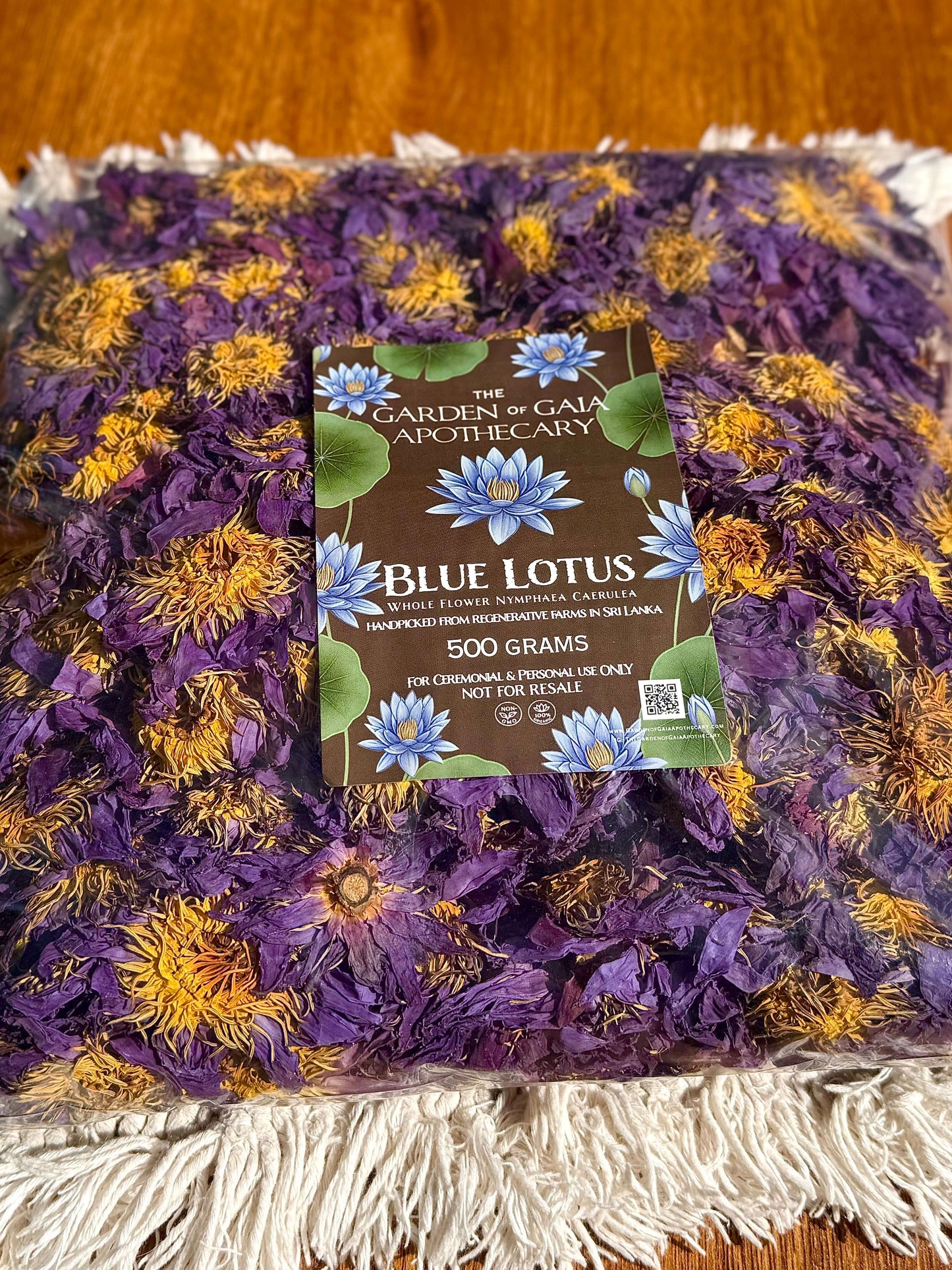 A 1 kilo, 500 gram bag of dried blue lotus flowers for wholesale from Garden of Gaia Apothecary, displayed on a white fringed blanket over a wooden surface. The dried flowers are a mix of purple, yellow, and green, with a brown paper label in the center.