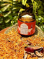 A jar of Hathor's Nectar, Saffron and frankincense tallow- front view. sits on a woven tray surrounded by dried herbs and botanicals, with a blurred green fern background.