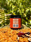 A jar of Hathor's Nectar, Saffron and frankincense tallow- side view. sits on a woven tray surrounded by dried herbs and botanicals, with a blurred green fern background.